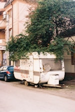 A weathered caravan parked on a street, partly covered by a large, leafy tree growing over it. There is graffiti on the side of the caravan, and a blue car parked next to it on the left. The area appears urban, with a multi-story building visible in the background.