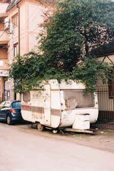 A weathered caravan parked on a street, partly covered by a large, leafy tree growing over it. There is graffiti on the side of the caravan, and a blue car parked next to it on the left. The area appears urban, with a multi-story building visible in the background.