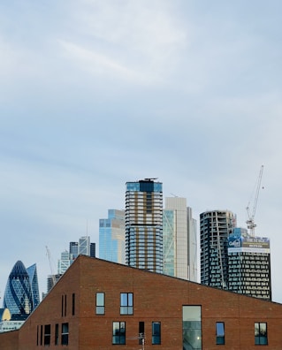 A cityscape featuring modern skyscrapers in the background and a brick building in the foreground. The skyscrapers include varied architectural styles, with sleek glass facades and some under construction. A crane is visible on the right side, indicating ongoing development.