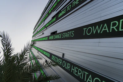 A modern building facade with multiple linear LED display panels, each showing different motivational and futuristic phrases in green text. A palm tree's leaves are visible in the foreground, adding a natural element to the scene.