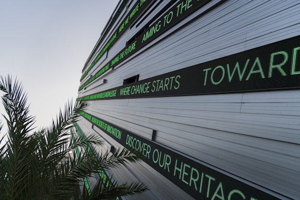 A modern building facade with multiple linear LED display panels, each showing different motivational and futuristic phrases in green text. A palm tree's leaves are visible in the foreground, adding a natural element to the scene.