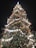 A large, beautifully decorated Christmas tree adorned with an array of glowing lights, colorful ornaments, and ribbons. The tree's decorations include red baubles, miniature houses, snowmen, and other festive items, all illuminated by the bright white lights that create a warm and inviting atmosphere against the dark night sky.