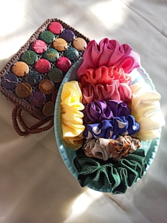 A collection of colorful scrunchies arranged in a light blue bowl, displaying shades of pink, red, yellow, purple, animal print, and green. Beside the bowl, there is a woven basket adorned with circular discs in various colors including green, pink, purple, and brown.