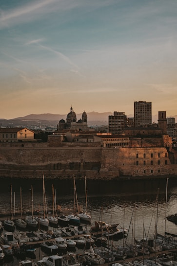 A coastal cityscape at dusk features a historic stone fortification in the foreground, with its warm tones contrasting against the darkening sky. The backdrop includes several prominent architectural structures, including a cathedral with domes and modern-looking buildings. Numerous boats are docked in a marina, their masts creating vertical lines against the tranquil water.