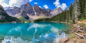 A breathtaking mountainous landscape featuring a pristine turquoise lake surrounded by dense evergreen forests. The towering rocky peaks are capped with snow, reflecting majestically in the calm water. Crystal-clear skies with scattered fluffy clouds enhance the serene and picturesque setting.