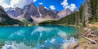A breathtaking mountainous landscape featuring a pristine turquoise lake surrounded by dense evergreen forests. The towering rocky peaks are capped with snow, reflecting majestically in the calm water. Crystal-clear skies with scattered fluffy clouds enhance the serene and picturesque setting.