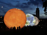 Large illuminated spheres representing planets are displayed outdoors at dusk. The foreground features a bright orange sphere resembling Mars, with Earth and the Moon visible beside it. Silhouetted people observe the spheres beneath a darkening sky, with a crescent moon visible in the sky and a tall building in the background.