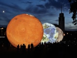 Large illuminated spheres representing planets are displayed outdoors at dusk. The foreground features a bright orange sphere resembling Mars, with Earth and the Moon visible beside it. Silhouetted people observe the spheres beneath a darkening sky, with a crescent moon visible in the sky and a tall building in the background.