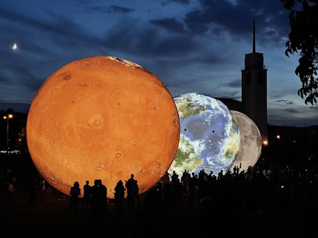 Large illuminated spheres representing planets are displayed outdoors at dusk. The foreground features a bright orange sphere resembling Mars, with Earth and the Moon visible beside it. Silhouetted people observe the spheres beneath a darkening sky, with a crescent moon visible in the sky and a tall building in the background.