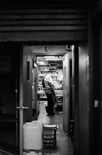 A person wearing a chef's uniform is standing in what appears to be a kitchen area. The scene is viewed through a doorway from a darker room or corridor. The overall ambiance suggests a busy and professional kitchen environment.