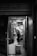 A person wearing a chef's uniform is standing in what appears to be a kitchen area. The scene is viewed through a doorway from a darker room or corridor. The overall ambiance suggests a busy and professional kitchen environment.