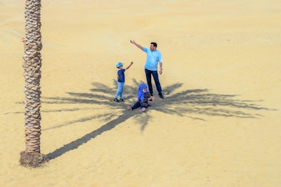 A family scene in a desert setting with a tall palm tree casting a shadow on the sand. A man, woman, and child are interacting near the tree. The man and child are standing, while the woman is seated, and they all appear to be enjoying the moment.