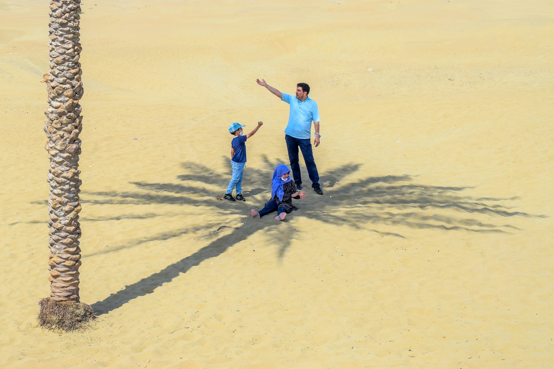 A family scene in a desert setting with a tall palm tree casting a shadow on the sand. A man, woman, and child are interacting near the tree. The man and child are standing, while the woman is seated, and they all appear to be enjoying the moment.
