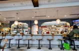 A cozy restaurant interior featuring a long counter with several empty bar stools. The counter is adorned with a patterned design and has overhead hanging lights. Behind the counter are shelves filled with various bottles and kitchen supplies. A person is seated at one end of the counter with a green bag by their side.