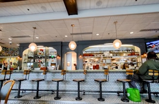 A cozy restaurant interior featuring a long counter with several empty bar stools. The counter is adorned with a patterned design and has overhead hanging lights. Behind the counter are shelves filled with various bottles and kitchen supplies. A person is seated at one end of the counter with a green bag by their side.