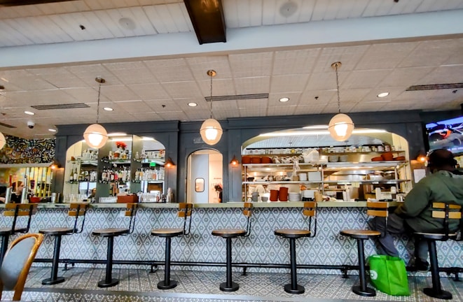 A cozy restaurant interior featuring a long counter with several empty bar stools. The counter is adorned with a patterned design and has overhead hanging lights. Behind the counter are shelves filled with various bottles and kitchen supplies. A person is seated at one end of the counter with a green bag by their side.
