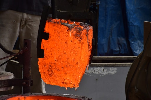 A glowing, molten metal crucible is being held by industrial tongs, with intense orange and red colors radiating from the liquid metal. The backdrop contrasts with shades of blue and gray, emphasizing the heat of the metal.