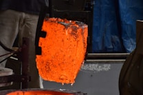 A glowing, molten metal crucible is being held by industrial tongs, with intense orange and red colors radiating from the liquid metal. The backdrop contrasts with shades of blue and gray, emphasizing the heat of the metal.