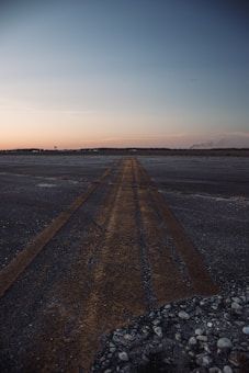 An expansive, empty runway stretches into the distance under a vast sky. The surface shows signs of wear with visible cracks and pebbles scattered across. The sky above transitions from a deep blue to a subtle peach hue on the horizon, indicating a sunrise or sunset.