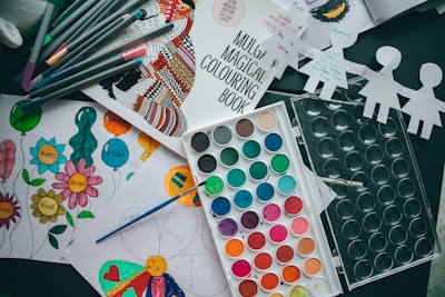 Various art supplies are scattered on a table, including a coloring book, colored markers, a watercolor paint palette, and several pieces of paper with drawings and colorings. The pieces of paper have images of balloons and flowers with emotions written on them, and some cut-out paper doll figures are seen as well.