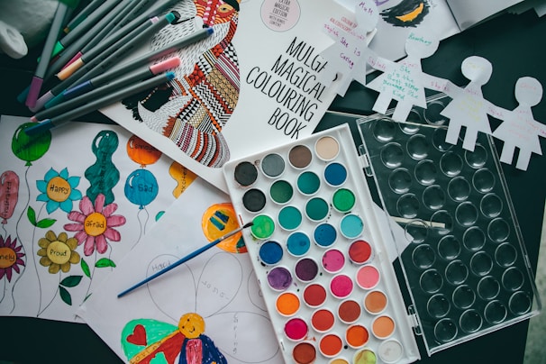 Various art supplies are scattered on a table, including a coloring book, colored markers, a watercolor paint palette, and several pieces of paper with drawings and colorings. The pieces of paper have images of balloons and flowers with emotions written on them, and some cut-out paper doll figures are seen as well.