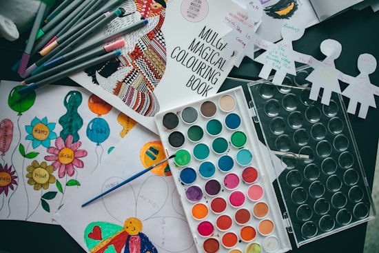 Various art supplies are scattered on a table, including a coloring book, colored markers, a watercolor paint palette, and several pieces of paper with drawings and colorings. The pieces of paper have images of balloons and flowers with emotions written on them, and some cut-out paper doll figures are seen as well.