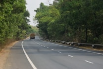A long, empty road stretches into the distance, flanked by lush green trees on both sides. A single large vehicle, possibly a truck, is visible in the distance moving away. The road appears to be well-maintained and lined with a metal barrier.