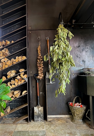 A rustic setting featuring a wall with various items hanging from it including a bunch of dried garlic and a large bundle of green leaves. There is a neatly arranged stack of firewood in a shelf nearby. Below, a shovel and a spade are leaning against the wall. A decorative metal basket filled with red objects, possibly tomatoes, is placed on the ground.