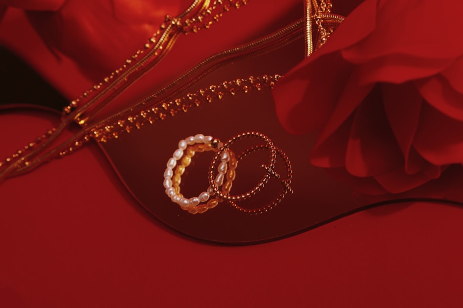 A collection of bracelets, including a pearl bracelet, and gold chain jewelry are arranged on a reflective surface. There is a rose in the image and a zipper that appears to belong to a bag or pouch, all set against a red background.