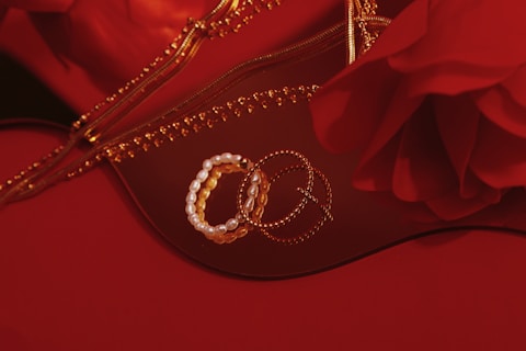 A collection of bracelets, including a pearl bracelet, and gold chain jewelry are arranged on a reflective surface. There is a rose in the image and a zipper that appears to belong to a bag or pouch, all set against a red background.