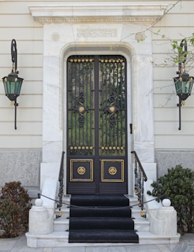 The image features an elegant entrance with a decorative black iron gate adorned with gold accents. The gate is set within a white marble frame, and there are ornamental black and gold lamps on either side. Steps lead up to the entrance, bordered by plants and shrubs, adding to the sophisticated appearance.