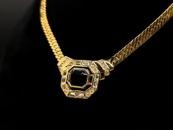A gold necklace with a thick chain features a large, central pendant. The pendant has an octagonal shape adorned with a prominent black gemstone in the center, surrounded by smaller clear stones set in a gold frame. The background is a deep black, highlighting the luxurious details of the necklace.