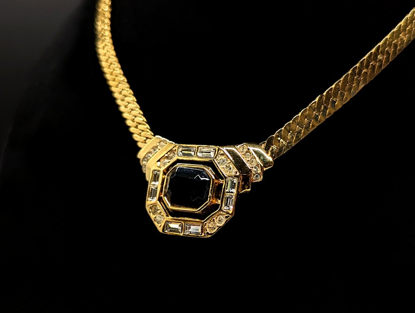 A gold necklace with a thick chain features a large, central pendant. The pendant has an octagonal shape adorned with a prominent black gemstone in the center, surrounded by smaller clear stones set in a gold frame. The background is a deep black, highlighting the luxurious details of the necklace.