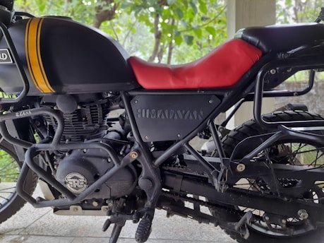 A close-up view of a motorcycle with a focus on its engine and frame. The motorcycle has a black body with yellow stripes on the fuel tank and a red seat. The brand name appears on the side panel. Details such as the engine components, chain, and tires are visible. The background shows a blurry outdoor setting with green foliage.