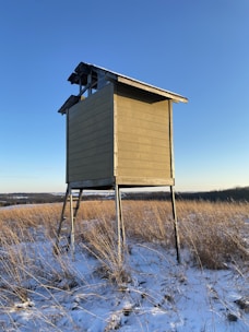 A solitary wooden hunting stand elevated on stilts sits in an open grassy field lightly covered in snow. The structure is weathered and stands against a clear blue sky, with the sun casting shadows on its surface.