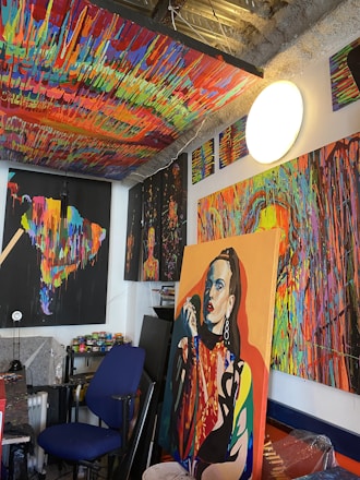 A vividly colored art studio is filled with multiple abstract paintings that feature bold and bright dripping paint patterns. There is a prominent portrait of a woman in one of the paintings. The studio has a cluttered yet creative atmosphere, with various art supplies scattered around, including paint cans and brushes. The ceiling is also painted with a vibrant, multicolored pattern.