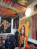 A vividly colored art studio is filled with multiple abstract paintings that feature bold and bright dripping paint patterns. There is a prominent portrait of a woman in one of the paintings. The studio has a cluttered yet creative atmosphere, with various art supplies scattered around, including paint cans and brushes. The ceiling is also painted with a vibrant, multicolored pattern.