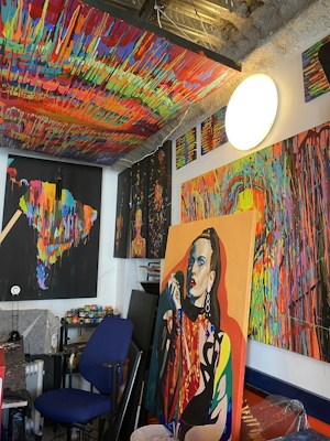A vividly colored art studio is filled with multiple abstract paintings that feature bold and bright dripping paint patterns. There is a prominent portrait of a woman in one of the paintings. The studio has a cluttered yet creative atmosphere, with various art supplies scattered around, including paint cans and brushes. The ceiling is also painted with a vibrant, multicolored pattern.