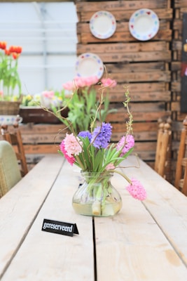 A wooden table features a glass vase with pink, purple, and blue flowers. A small sign with the word 'gereserveerd' rests on the table, indicating a reserved spot. The background includes rustic wooden crates and decorative plates mounted on a wall. There are additional flowers in the background, with soft natural lighting enhancing the cozy atmosphere.