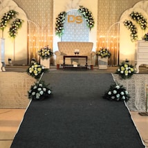 An elegant stage setup with floral decorations arranged symmetrically around a beige couch. Soft lighting creates a warm ambiance, complemented by a patterned backdrop with initials 'DS'. Flowers in white and yellow are arranged in bouquets on either side and at the base of the stage, with decorative arches flanking the setup.