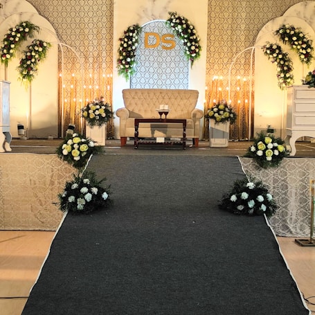 An elegant stage setup with floral decorations arranged symmetrically around a beige couch. Soft lighting creates a warm ambiance, complemented by a patterned backdrop with initials 'DS'. Flowers in white and yellow are arranged in bouquets on either side and at the base of the stage, with decorative arches flanking the setup.