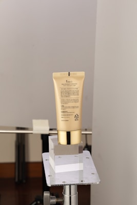 A beige cosmetic tube with gold accents is placed on a metallic stand. The tube features text and branding and appears to be a skincare product. A minimalistic and clean background is used, primarily showcasing the product without distractions.