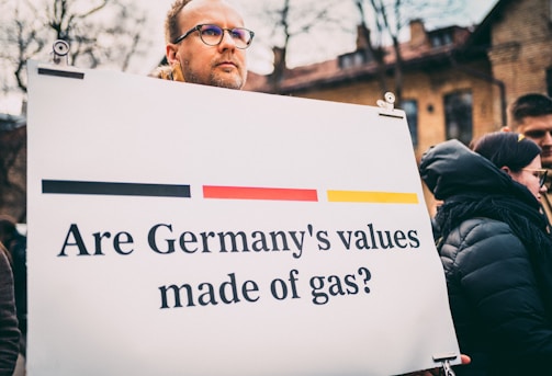A person holding a sign with the text 'Are Germany's values made of gas?' The setting appears to be outdoors with several people in the background. The sign includes horizontal stripes in the colors black, red, and yellow, reminiscent of the German flag.
