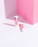 Two tubes of lip balm with pink, red, and blue packaging are placed on a light pink and white geometric background. The tubes cast soft shadows, adding depth to the minimalist setting.
