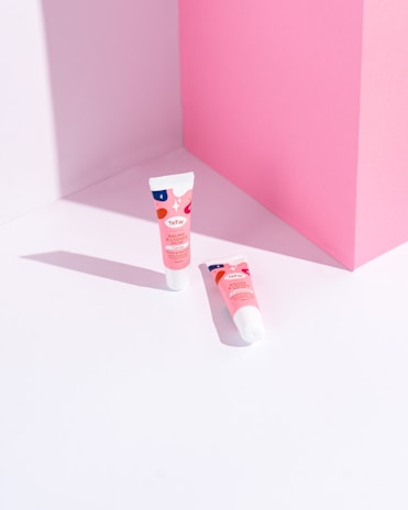 Two tubes of lip balm with pink, red, and blue packaging are placed on a light pink and white geometric background. The tubes cast soft shadows, adding depth to the minimalist setting.