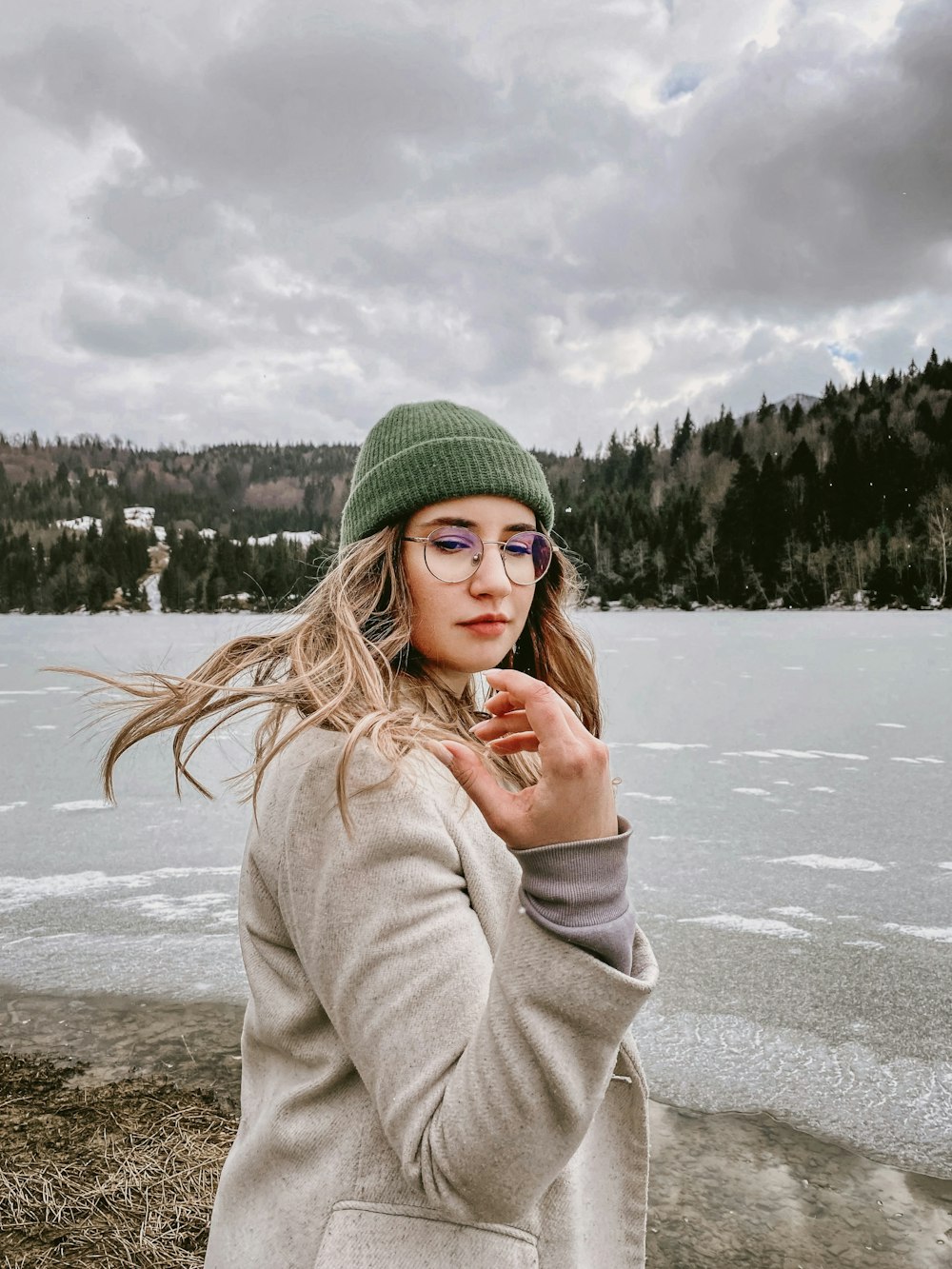 a woman wearing a hat and glasses standing in front of a lake