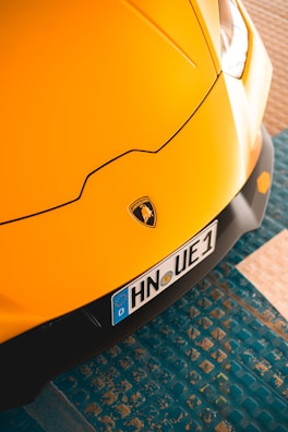 A close-up view of a yellow sports car's front, featuring the Lamborghini logo prominently on the hood. The car's design lines are sleek and emphasize aerodynamic aesthetics. The license plate with the registration number is visible, set against a textured blue floor.