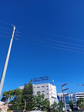 A multistory medical center building with a sign that reads 'Mary Mediatrix Medical Center' is surrounded by tall green trees. In the foreground, there are power lines and a tall utility pole. The sky is clear and bright blue.