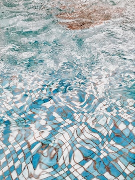 Rippling water creates a wavy pattern over a blue and white tiled swimming pool floor. The distortion of the tiles by the water's surface tension produces an abstract, fluid design.