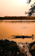 A serene river scene at sunset with a silhouette of a canoe and a person sitting inside. The water reflects the warm hues of the sky, creating a tranquil ambiance. Trees and foliage are visible in the foreground and background, enhancing the natural setting.